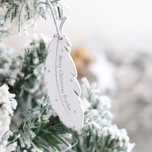 A close-up of a Christmas tree branch adorned with a sentimental touch—a Christmas Family Sentimental Feather Hanger. The white, feather-shaped ornament is inscribed with "Merry Christmas, Brother" and is beautifully surrounded by frosty, snow-dusted greenery.