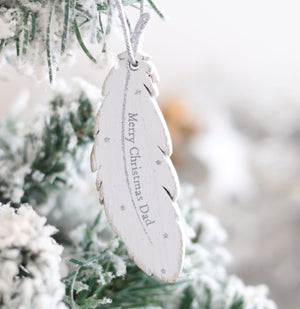 A close-up of the Christmas Family Sentimental Feather Hanger ornament, gracefully hanging on a snow-covered tree. Inscribed with "Merry Christmas Dad" in a delicate font and surrounded by small stars, it evokes cherished memories of Christmas family gatherings.