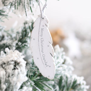 A Christmas Family Sentimental Feather Hanger adorns a snow-covered tree branch. The cherished memory features the engraved message, "Merry Christmas Grandpa!" Small silver stars decorate the feather, adding a festive touch to the wintry scene.