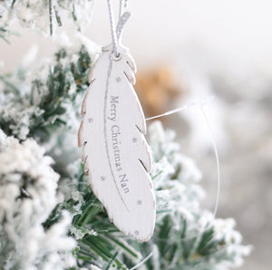 A close-up of the Christmas Family Sentimental Feather Hanger, a feather-shaped ornament with "Merry Christmas Nan" written on it. Hanging on a frosted, snow-covered branch of an artificial tree, this cherished hanger adds a sentimental touch to our Christmas family collection.