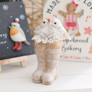 Product Description: A charming figurine featuring two white birds nestled inside a pair of textured brown wellies adorned with floral accents, elegantly displayed on a light surface. In the softly blurred background, festive signs showcase illustrations of birds and gingerbread figures—an ideal addition to your Christmas Birds Sitting On Wellies Ornament collection.