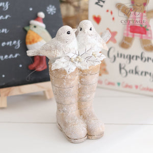 The Christmas Birds Sitting On Wellies Ornament features two white bird figurines perched on vintage-style wellies, which are beautifully adorned with white flowers and berries. The boots are dusted with a snowy effect, adding to their charm. In the background, festive decorations include signs with gingerbread themes.