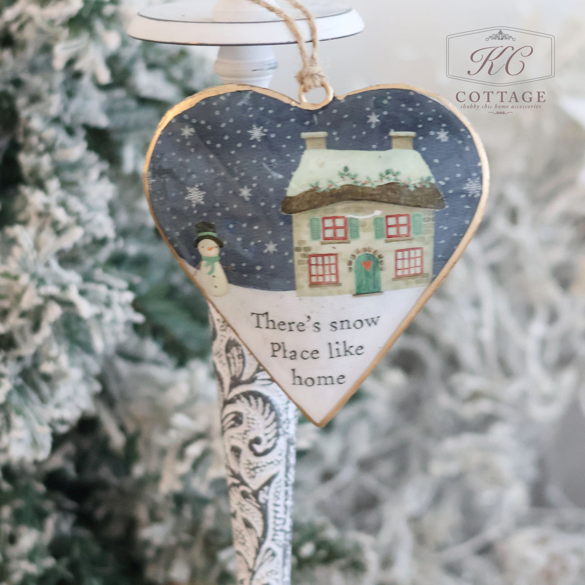 The Christmas Metal Cottage Home Hanging Hearts ornament showcases a snowy cottage scene with a snowman and the phrase "There's snow place like home." This delightful Christmas decoration is elegantly displayed hanging from a decorative stand in front of a frosted Christmas tree.