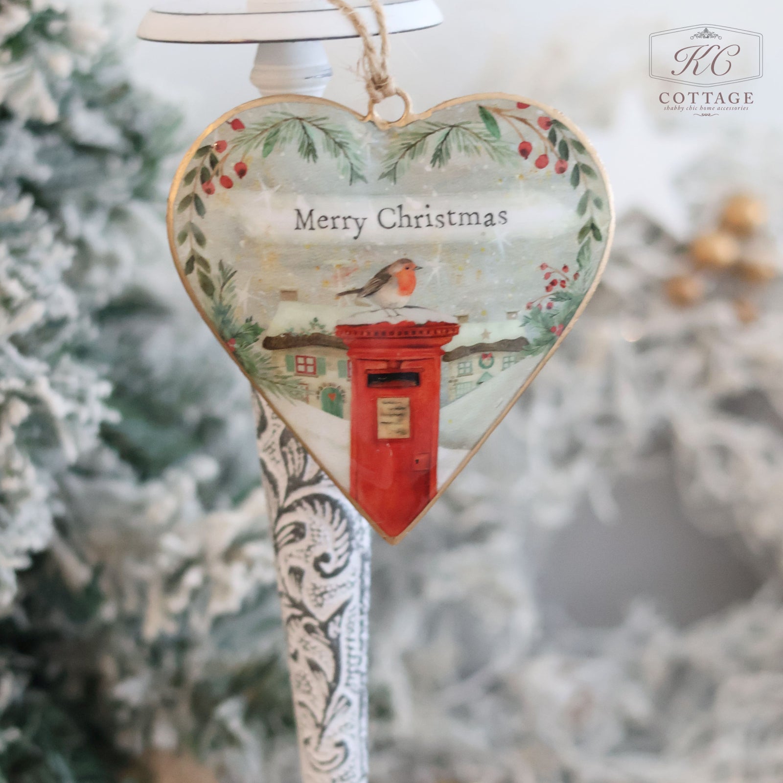 Christmas Metal Cottage Home Hanging Heart ornament featuring a robin perched on a red postbox with "Merry Christmas" text. This delightful piece of holiday decor is embellished with holly and greenery accents, set against a blurred winter background evocative of snowy cottage scenes.