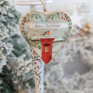 Christmas Metal Cottage Home Hanging Heart ornament featuring a robin perched on a red postbox with "Merry Christmas" text. This delightful piece of holiday decor is embellished with holly and greenery accents, set against a blurred winter background evocative of snowy cottage scenes.