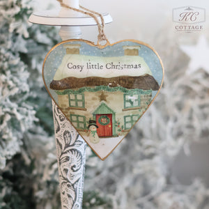 Hanging amidst the frosted greenery is a heart-shaped ornament from the "Christmas Metal Cottage Home Hanging Hearts" collection. It features an enchanting snowy cottage scene with a red door and the inscription "Cosy little Christmas" above it, adding charm to the festive decor.