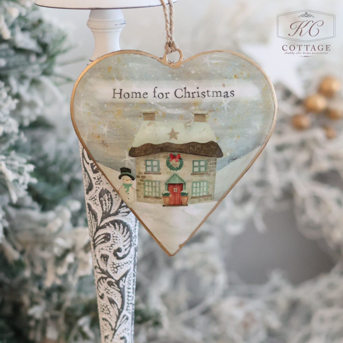 The Christmas Metal Cottage Home Hanging Hearts ornament showcases a snowy cottage scene with the message "Home for Christmas." This charming piece, adorned with a snowman and surrounded by festive decor, adds character to your hanging hearts collection against a frosted backdrop.
