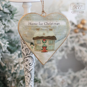 The Christmas Metal Cottage Home Hanging Hearts ornament showcases a snowy cottage scene with the message "Home for Christmas." This charming piece, adorned with a snowman and surrounded by festive decor, adds character to your hanging hearts collection against a frosted backdrop.