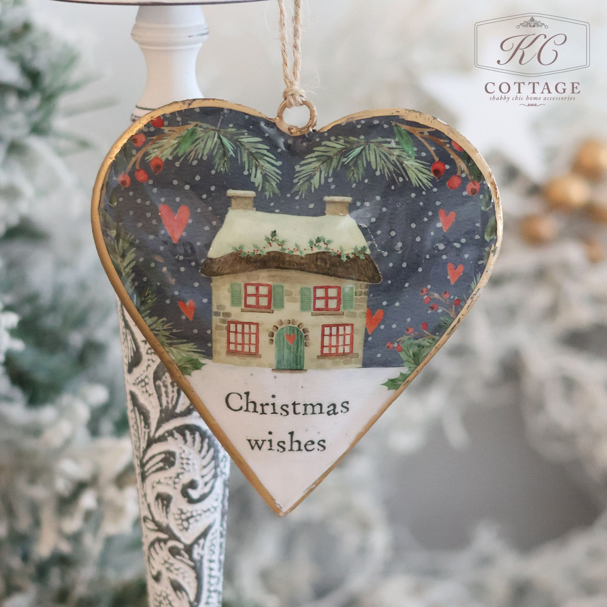 The Christmas Metal Cottage Home Hanging Hearts ornament showcases a heart-shaped design with a picturesque snowy cottage scene, complemented by hanging red hearts and green pine branches. The text "Christmas wishes" adds a festive touch. Set against a backdrop of a decorative stand and blurred greenery, it is ideal for enhancing your Christmas decor.