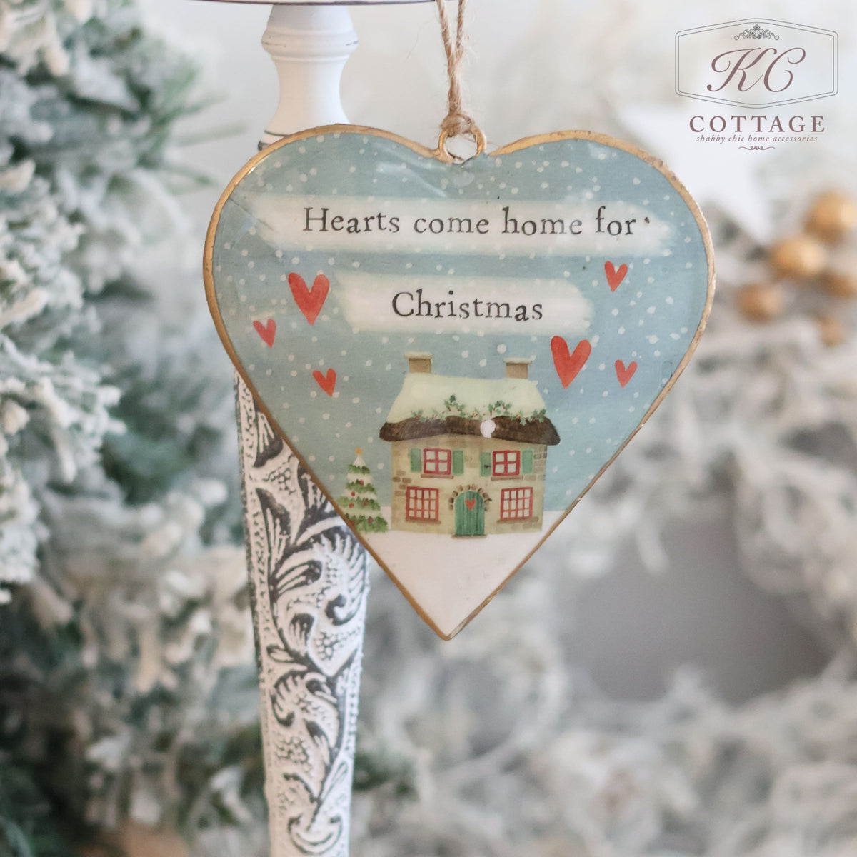 The Christmas Metal Cottage Home Hanging Hearts ornament is heart-shaped and features a snowy cottage scene with the phrase "Hearts come home for Christmas" alongside red hanging hearts. It dangles gracefully in front of a decorated tree, with the "KC Cottage" logo elegantly positioned in the top right corner.