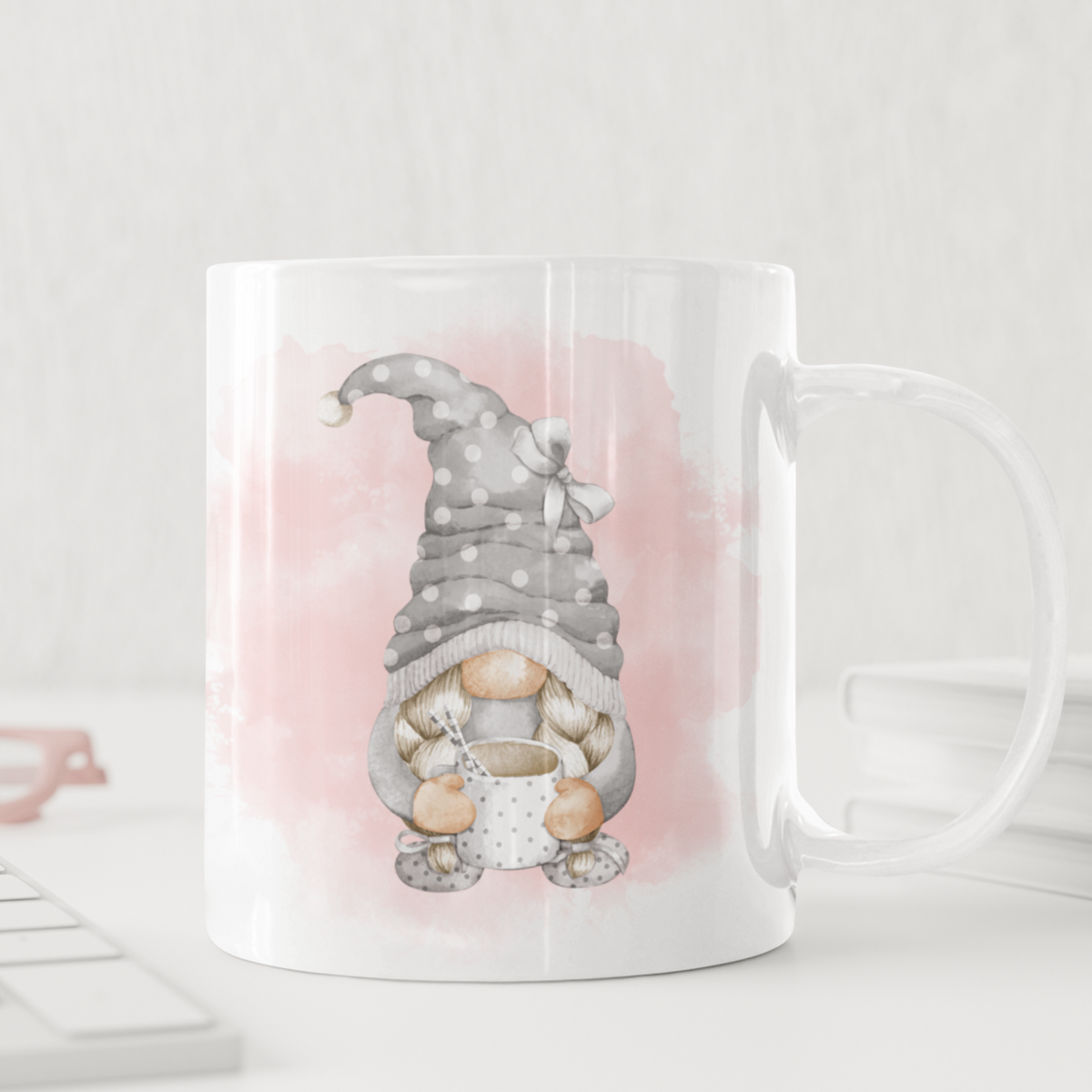 A Pink and Grey Gonk Mug with an illustration of a gnome in a gray, polka-dotted hat holding a cup of coffee. The light pink watercolor background adds charm, making it a delightful piece of home decor. The mug sits on a white desk surrounded by a keyboard, glasses, and stacked books.