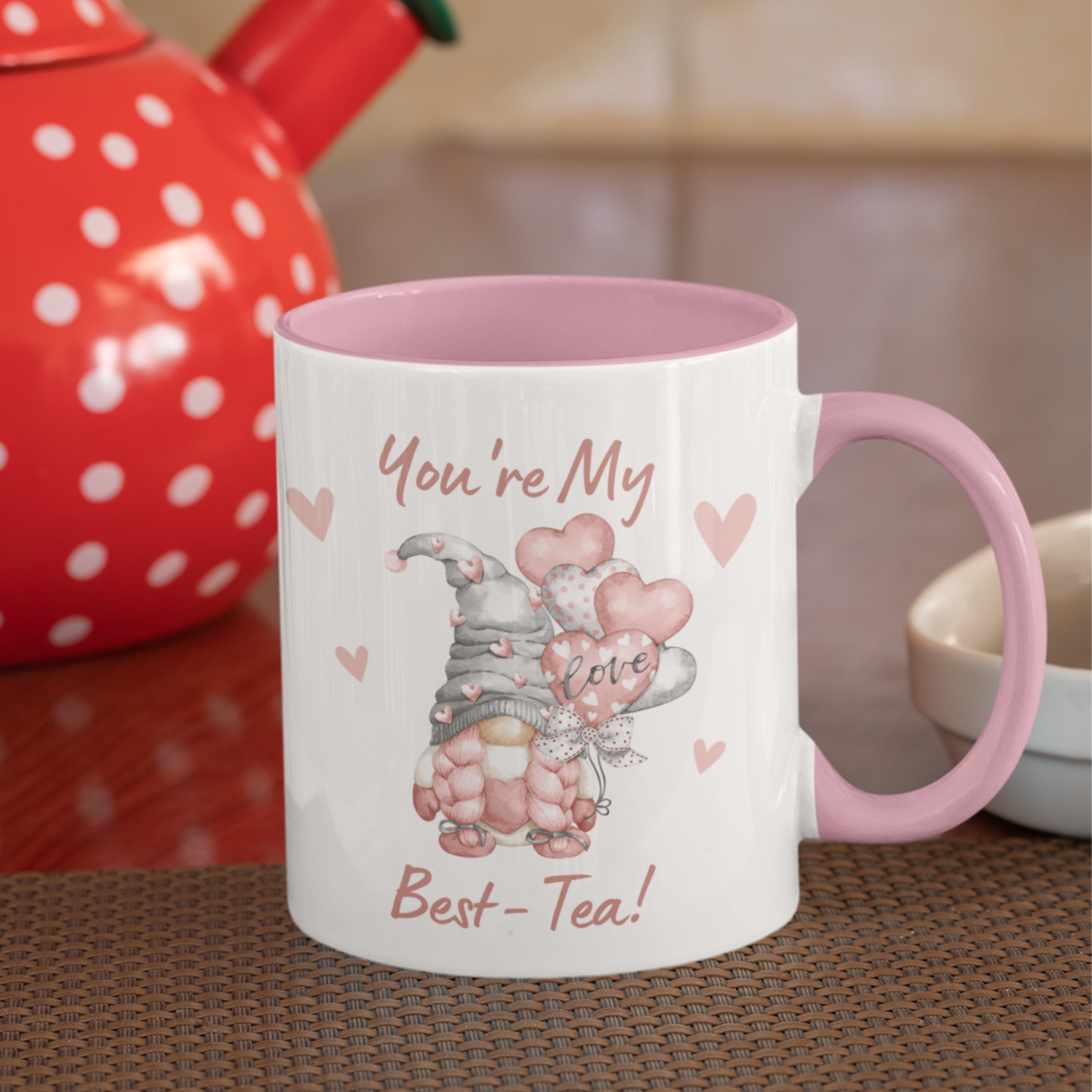 A white mug with a pink handle and interior features a cute gnome holding heart-shaped balloons. The text reads "You're My Best-Tea!" with "love" written on a balloon. Behind, there's a red teapot with white polka dots on a wooden table, adding a charming touch to your home decor. This adorable piece is the You're My Best-tea Gonk Mug.