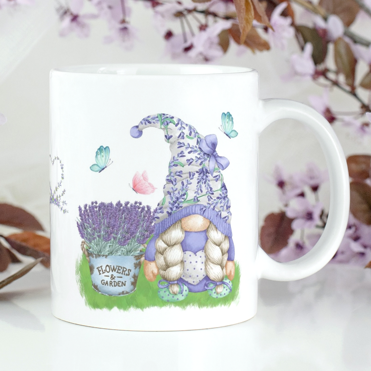 A white Gonk Lavender & Butterfly Mug is decorated with an illustration of a gnome wearing a lavender-patterned hat, next to a pot of lavender flowers labeled "Flowers & Garden." Butterflies hover around, and a pastel background with flowering branches complements the home decor scene.