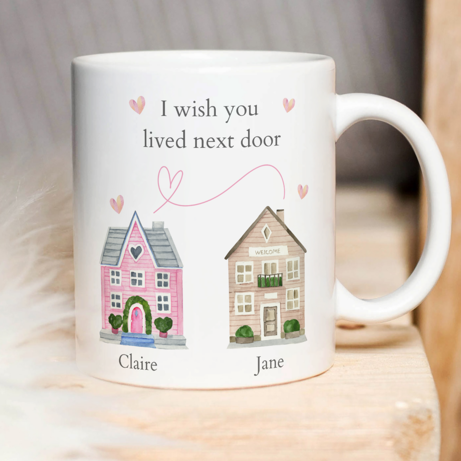 A I Wish You Lived Next Door Personalised Mug with two illustrated houses labeled "Claire" and "Jane" under the message "I wish you lived next door." One house is pink with heart details, and the other is beige with a "Welcome" sign. Pink hearts float between the two houses, making it a charming piece of home decor.