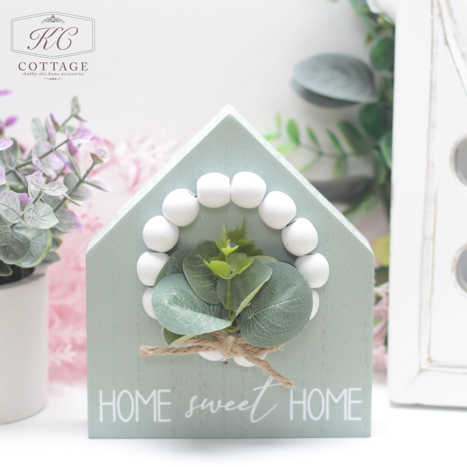 A small Wooden Block House with Bead & Foliage, adorned with a natural twine and leaf arrangement and a white bead wreath. The words "Home sweet Home" are printed at the bottom. Nearby, a potted purple plant and greenery add a cozy touch to your home decor.