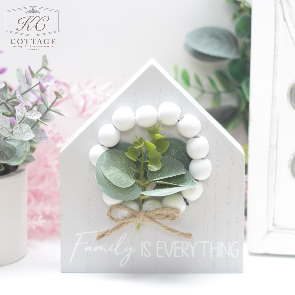 A small, house-shaped home decor piece features a wreath of white beads and green foliage on its front. The text "Family is Everything" is written below the wreath. The background showcases pink and green plants and part of a white decor item. The KC Cottage logo is in the top left. This Wooden Block House with Bead & Foliage makes for a charming addition to any living space.