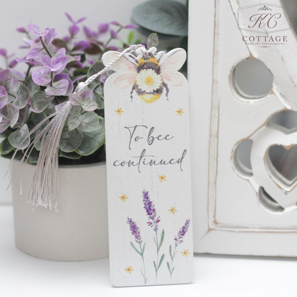 A Daisy Bee Bookmark with a tassel features a painted bee at the top, the text "To bee continued" in the middle, and lavender flowers at the bottom. Perfect for home decor, it is placed against a backdrop of purple flowers and a decorative white lantern.