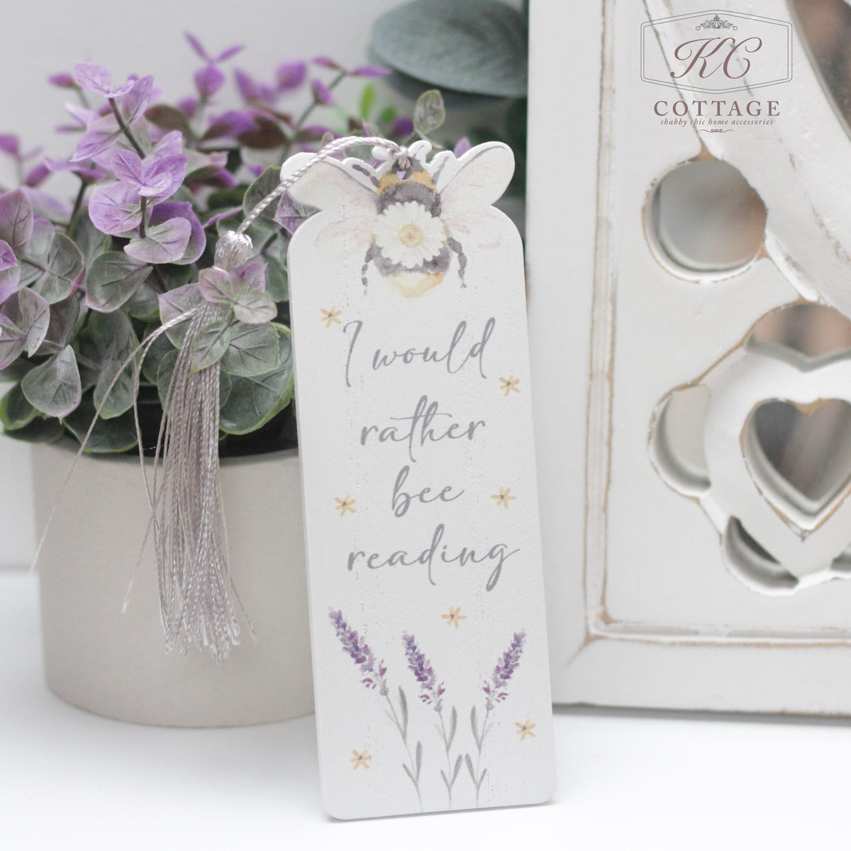 A white bookmark with a tassel displays the text "I would rather bee reading" accompanied by a bee illustration and lavender flowers. Set against a backdrop of purple blooms and decorative heart-shaped cutouts, this charming Daisy Bee Bookmarks piece doubles as delightful home decor.
