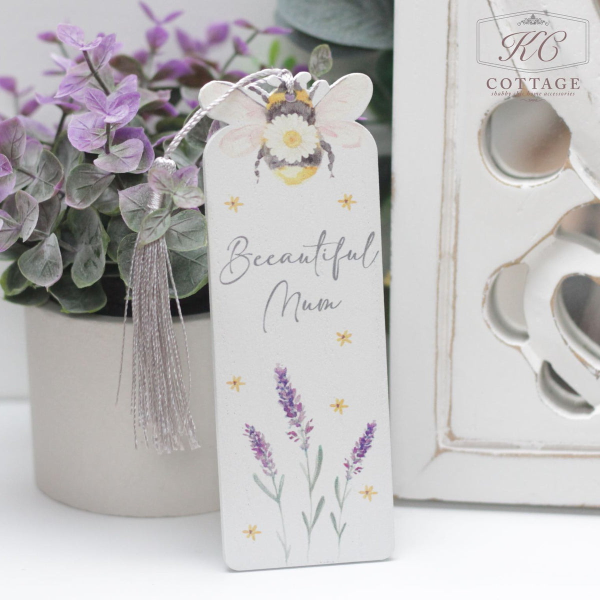 A decorative Daisy Bee Bookmarks with "Beeautiful Mum" written on it, adorned with illustrations of a bee, yellow flowers, and lavender flowers. It has a grey tassel attached to the top. Perfect for adding charm to your home decor, the bookmark is placed next to a small purple flower arrangement and a decorative white object.
