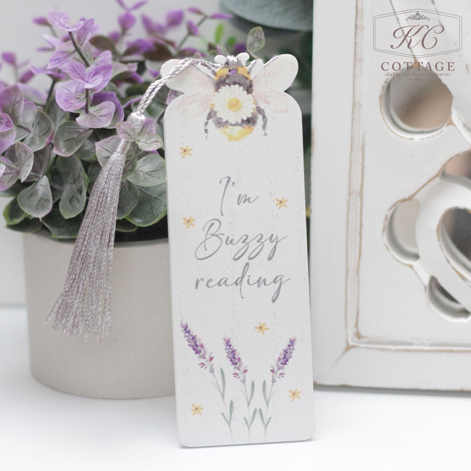 A Daisy Bee Bookmark stands upright, adorned with a bee illustration at the top and lavender flowers at the bottom. The text on the bookmark reads, "I'm buzzy reading." It is placed in a cozy home decor setting with potted plants and a white, ornate decor piece in the background.