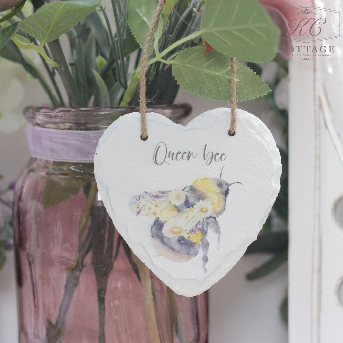 A Bee Slate Hanging Hearts with an illustration of a bee and the text "Queen Bee" hangs by a string in front of a glass jar containing a bouquet with purple and white flowers. The jar, tied with a purple ribbon, adds charm to your home decor. The background features leaves and part of a white structure.