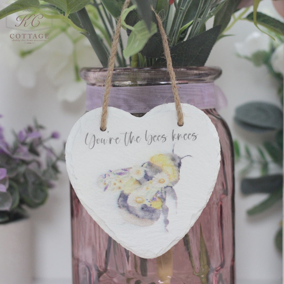 A Bee Slate Hanging Heart with an illustration of a bee and flowers hangs from a twine string. The text on the heart reads "You're the bees knees." This charming piece of home decor is attached to a pink glass vase containing various flowers and greenery.