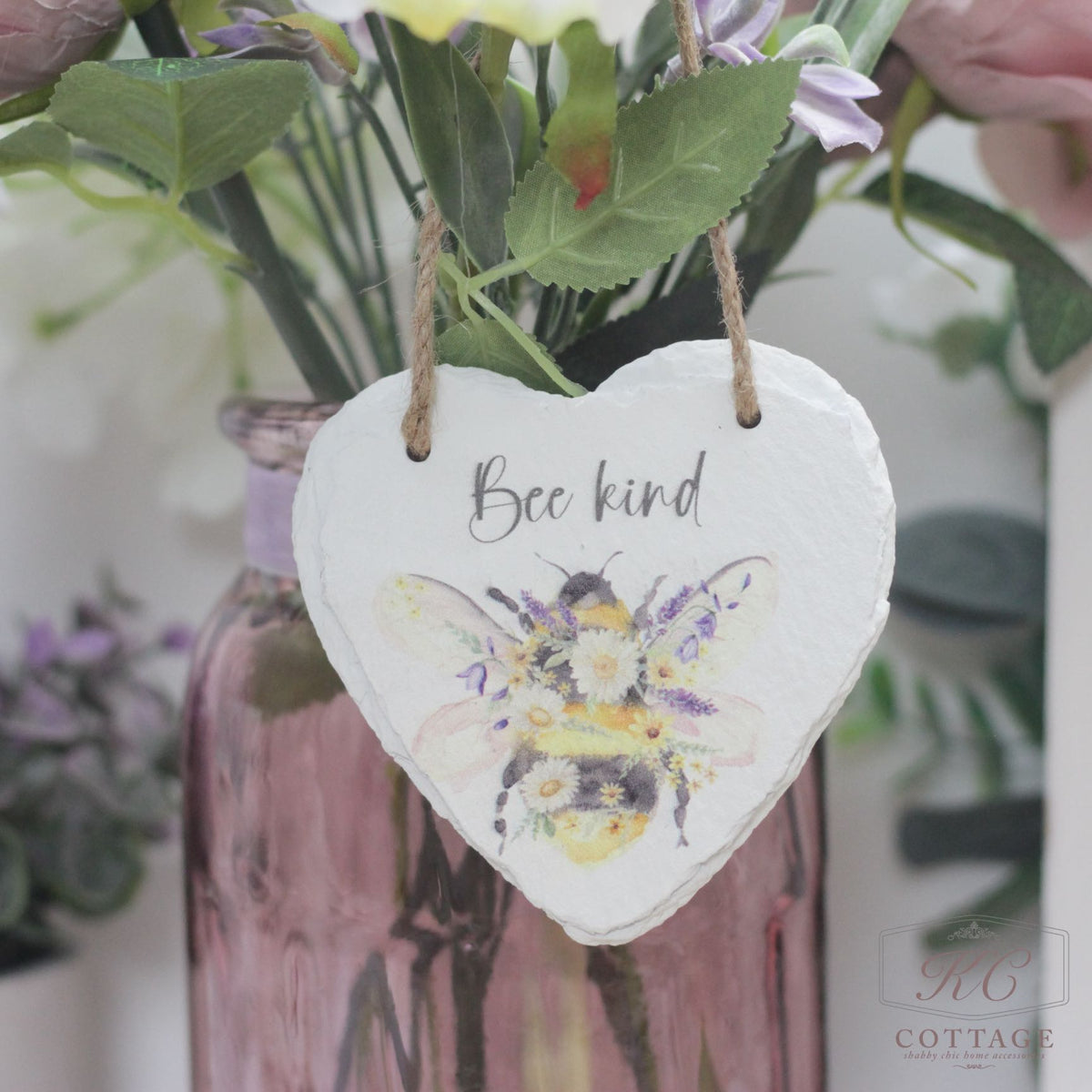 A Bee Slate Hanging Hearts sign with "Bee kind" written on it. The sign features a watercolor illustration of a bee and flowers, perfect for home decor. It's hanging by a twine cord in front of a mauve glass vase filled with flowers. The lower right corner shows a small logo: "KC Cottage.
