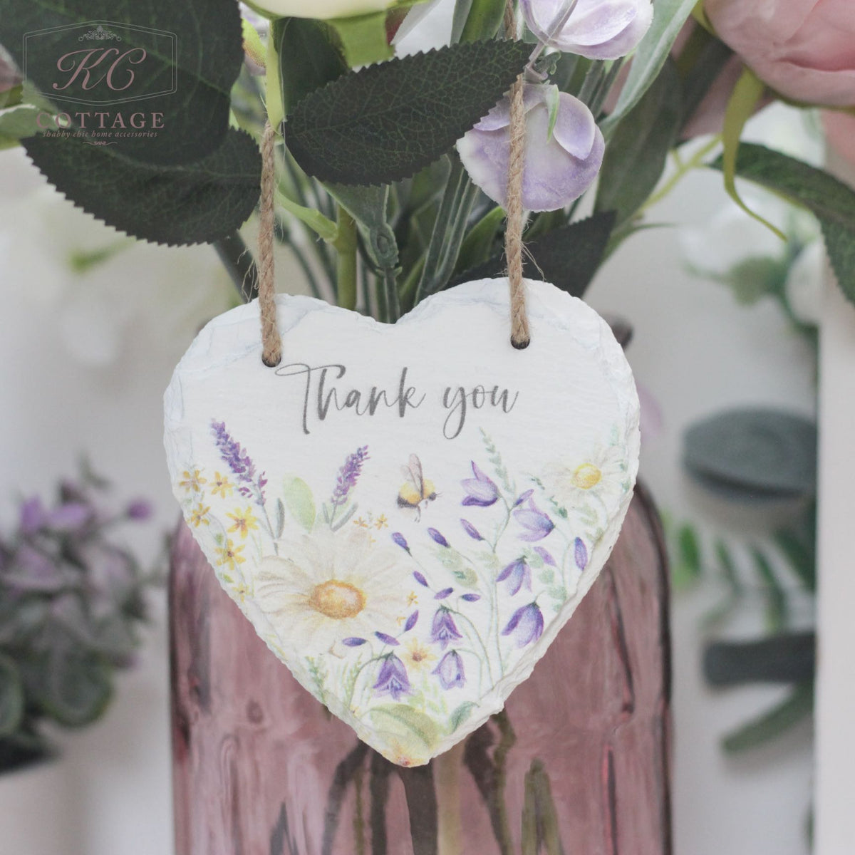 A Bee Slate Hanging Hearts made of white slate, adorned with colorful floral illustrations and the words "Thank you" written in the center, hangs from twine in front of a pink vase filled with purple and white flowers. This charming piece of home decor is set against a background of leaves and blurred flowers.