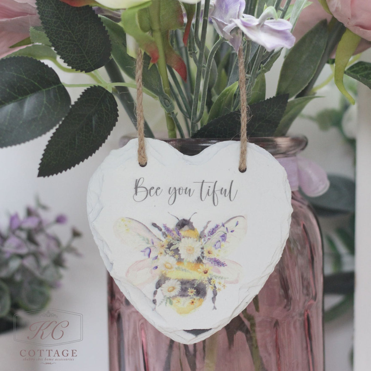 A decorative heart-shaped slate hangs by twine on a bouquet of flowers in a pink glass vase. The Bee Slate Hanging Hearts features an illustrated bee adorned with flowers and the text "Bee you tiful." The background includes leaves and flowers, adding a charming, rustic feel to this delightful piece of home decor.
