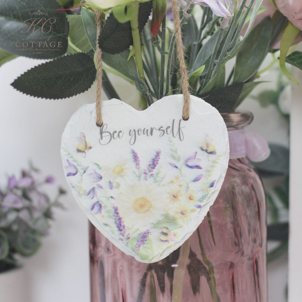 A Bee Slate Hanging Hearts sign with a watercolor floral design and the text "Bee yourself" hangs by a string in front of a bouquet of flowers in a pink vase. This charming piece of home decor is set against a background that includes more flowers and greenery, creating a soft, natural ambiance.