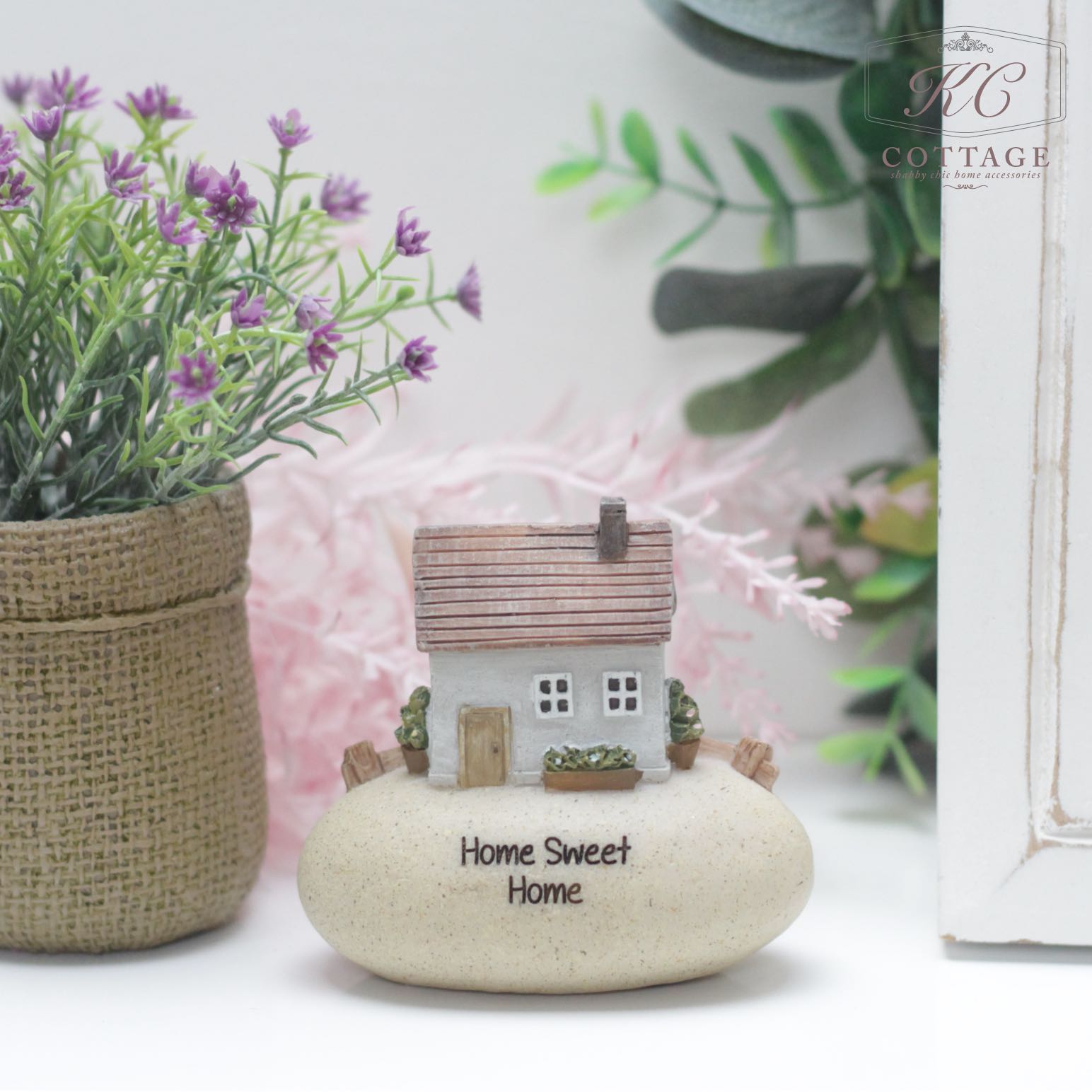 A small decorative model of a house with the words "Home Sweet Home" sits atop a smooth beige stone. The Pebble Lane Cottage Grey - Home Sweet Home features a grey house with a brown roof, surrounded by small green plants. In the background, there's a potted plant with purple flowers and lush foliage.
