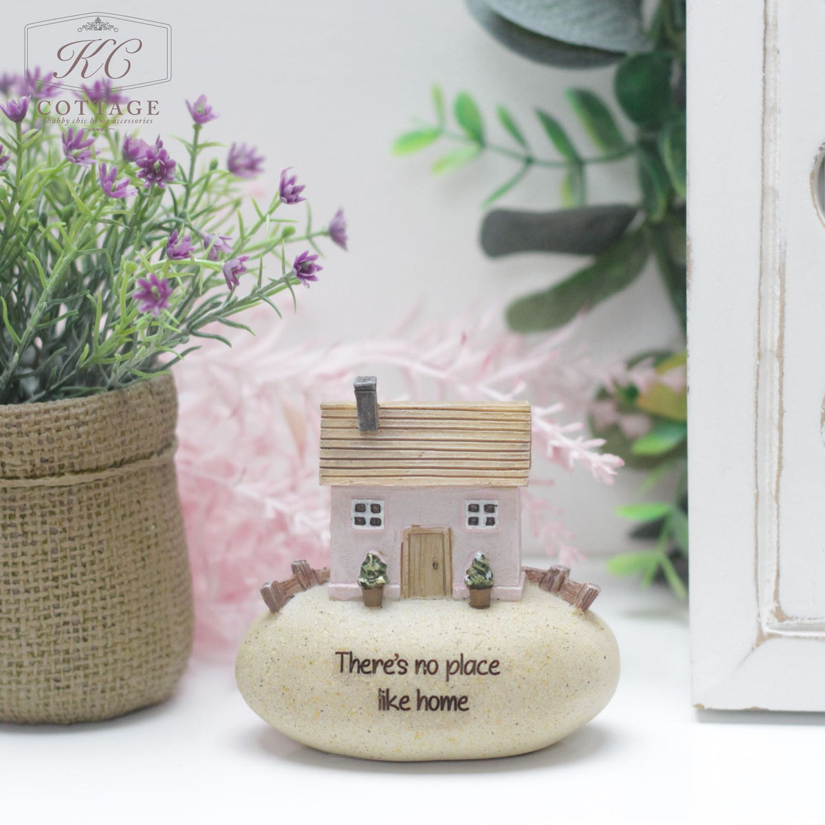 A miniature house model with a pink roof, Pebble Lane Cottage Pink - Theres No Place Like Home, sits on a smooth white stone inscribed with "There's no place like home." The home decor is placed on a white surface, accompanied by a potted plant featuring purple flowers and green foliage. The background includes greenery and a white cabinet.