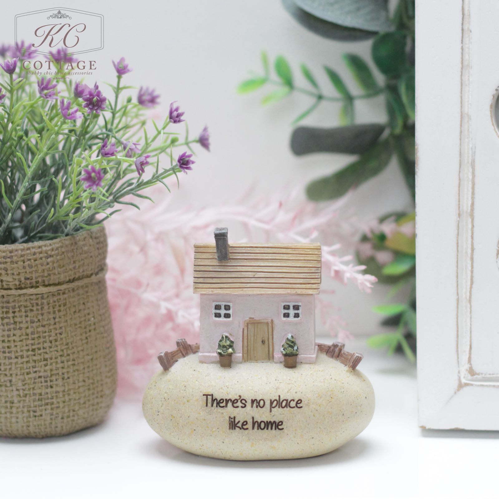 A miniature house model with a pink roof, Pebble Lane Cottage Pink - Theres No Place Like Home, sits on a smooth white stone inscribed with "There's no place like home." The home decor is placed on a white surface, accompanied by a potted plant featuring purple flowers and green foliage. The background includes greenery and a white cabinet.