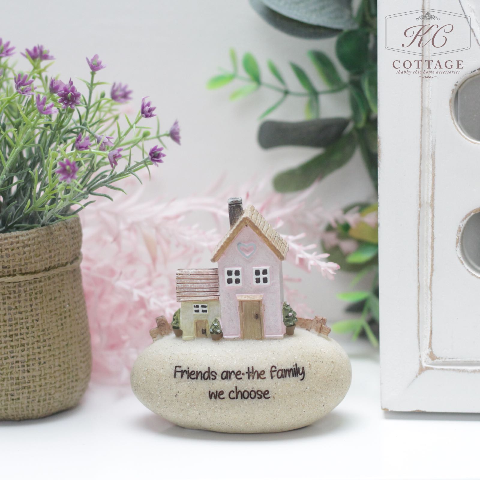 The Pebble Lane Cottage - Friends are the Family we Choose ornament showcases two miniature houses seated on a smooth, tan stone inscribed with "Friends are the Family we choose." Accompanying this charming scene is a potted plant adorned with purple flowers on the left side and partially visible wooden décor featuring small circular windows on the right.