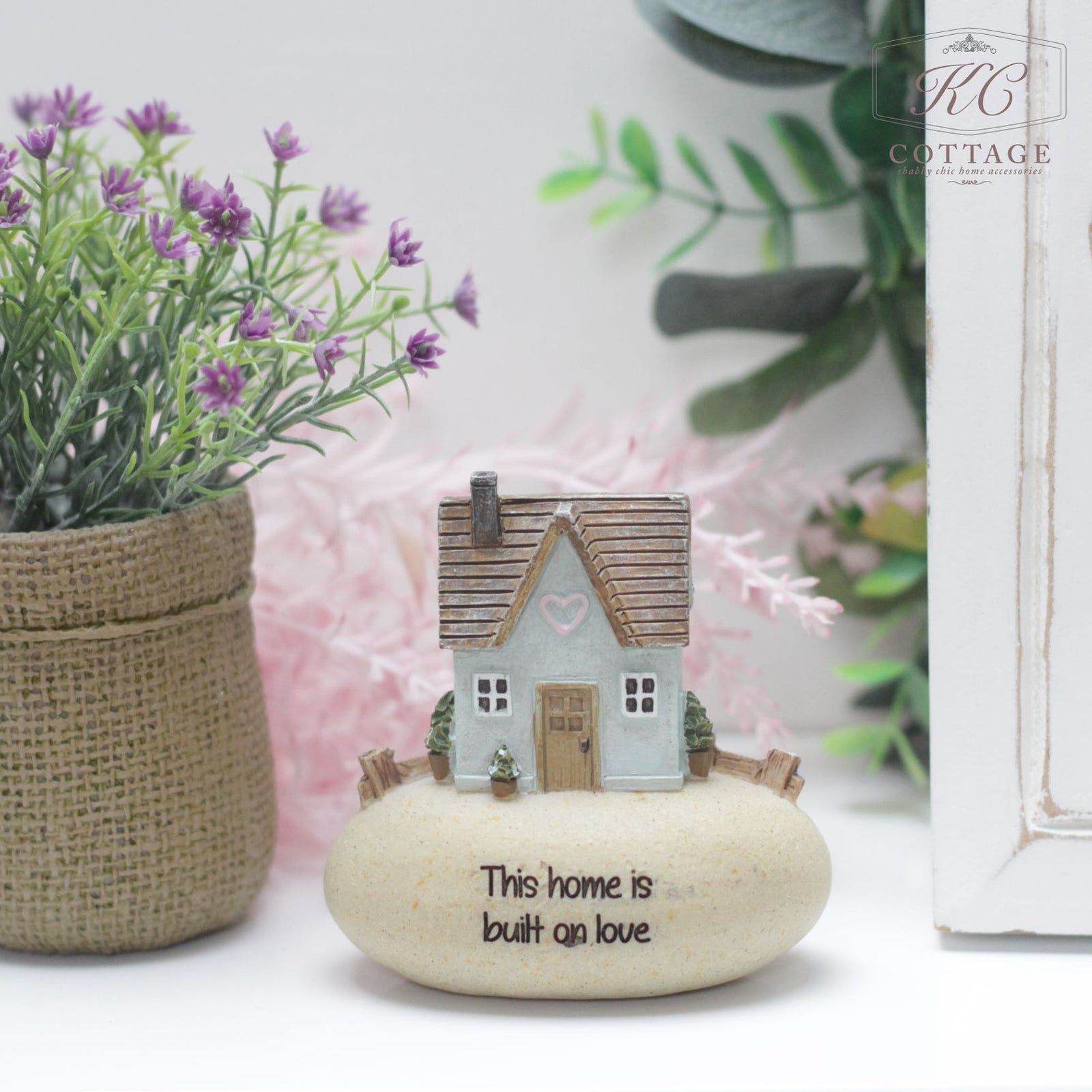 A small decorative home sits atop a round stone inscribed with "This Home Is Built On Love." The house is detailed with a heart above its door. Surrounding the stone is a potted plant with purple flowers to the left and greenery to the right, making it a charming piece of home decor. Introducing Pebble Lane Cottage Blue - This Home Is Built On Love, it’s the perfect addition to any household.