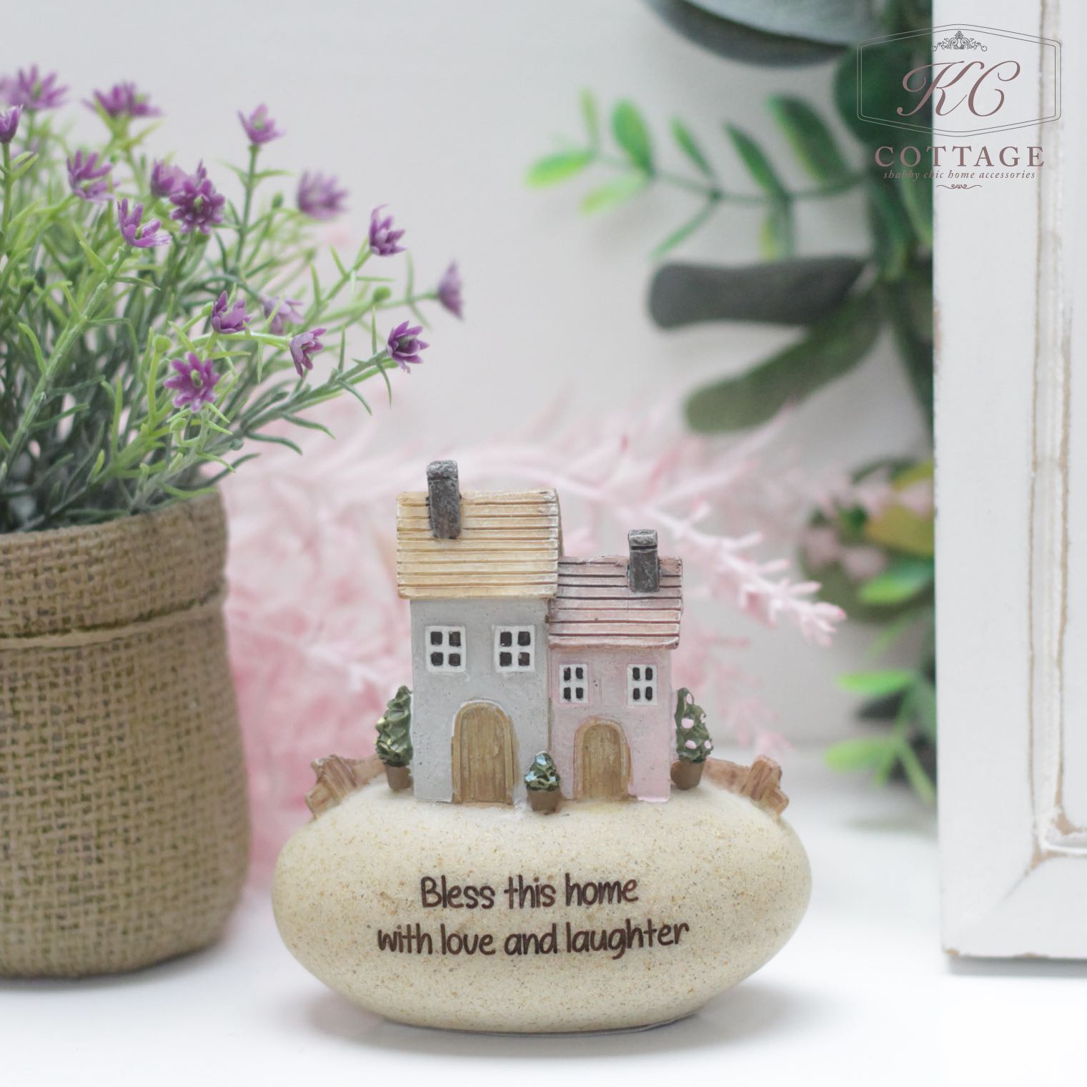 This Pebble Lane Cottage - Bless this home with love and laughter features two miniature houses and the text "Bless this home with love and laughter." Set on a white surface next to a potted plant with purple flowers and green leaves, it is accompanied by another plant in the background alongside a wooden frame.