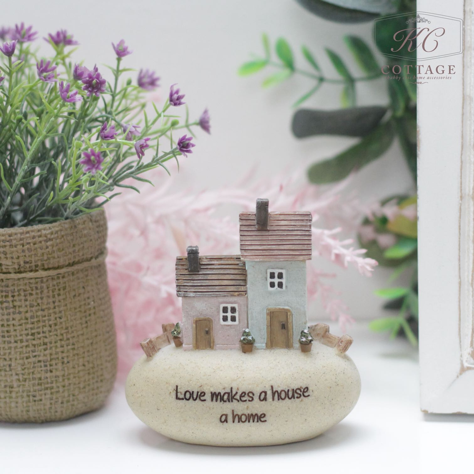 The Pebble Lane Cottage - Love makes a house a home ornament showcases two miniature houses perched on a smooth, oval rock featuring the heartfelt inscription "Love makes a house a home." The scene is beautifully accented with potted plants displaying lush green leaves and vibrant purple flowers, adding a touch of natural beauty.
