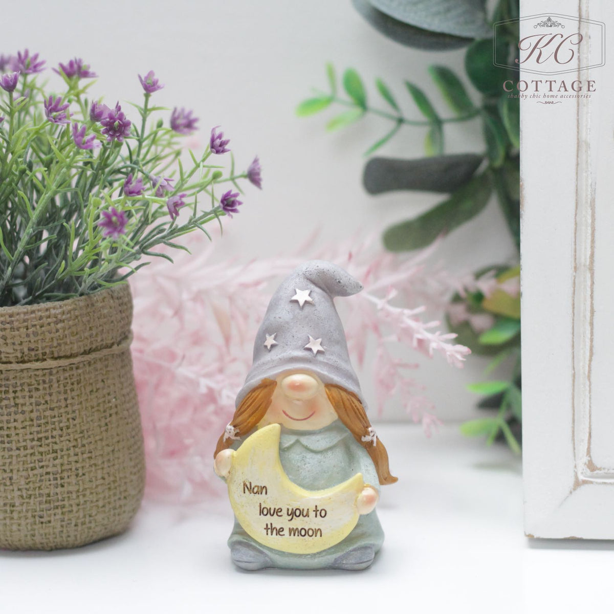 A small figurine of a smiling gnome with brown braided hair and a pointed gray hat featuring silver stars. The gnome holds a yellow crescent moon with the text "Nan love you to the moon." The background features a potted plant with purple flowers, making it perfect for home decor: the Nan Love you to the moon Gonk Ornament.