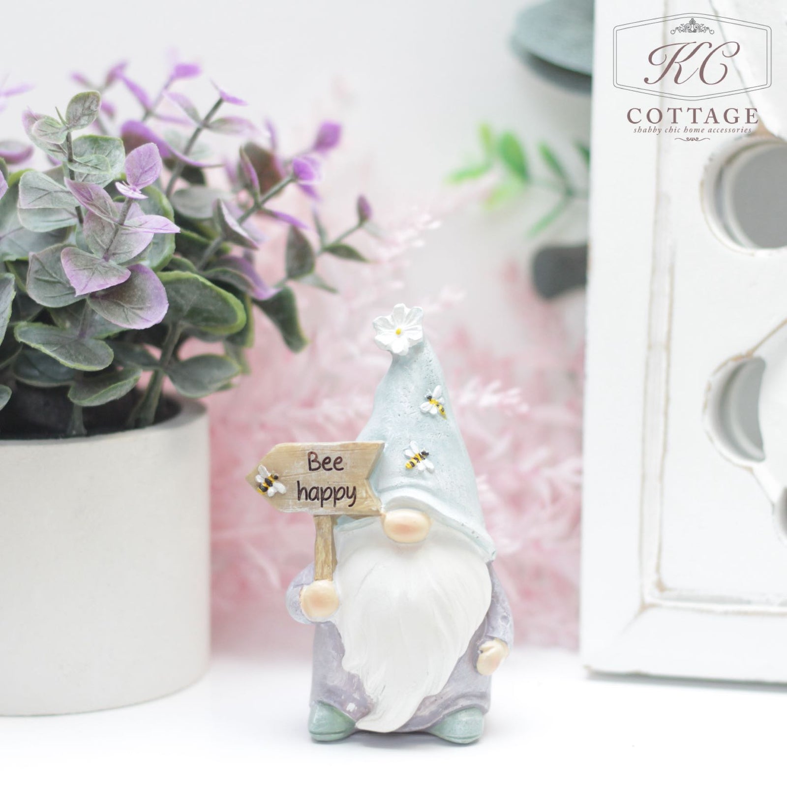 A small garden gnome with a white beard holds a sign that reads "Bee happy." The gnome, known as the Bee Happy Gonk Ornament, wears a light blue cone hat adorned with white flowers and bees. Surrounded by pink and green foliage, it's the perfect gift. The KC Cottage logo is visible in the top right corner.