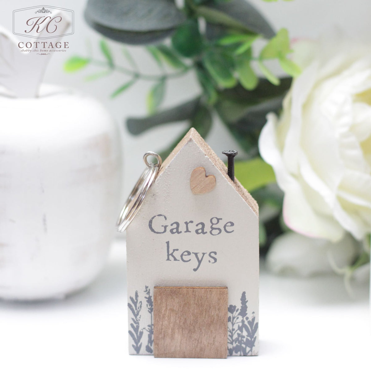 A small Chunky House Wooden Keyring in the shape of a house with the words "Garage keys" printed on it. Made of wood and painted white, it's adorned with a small wooden heart and a floral design at the bottom. Perfect for home decor, it sits in front of a white vase and flower in the background.