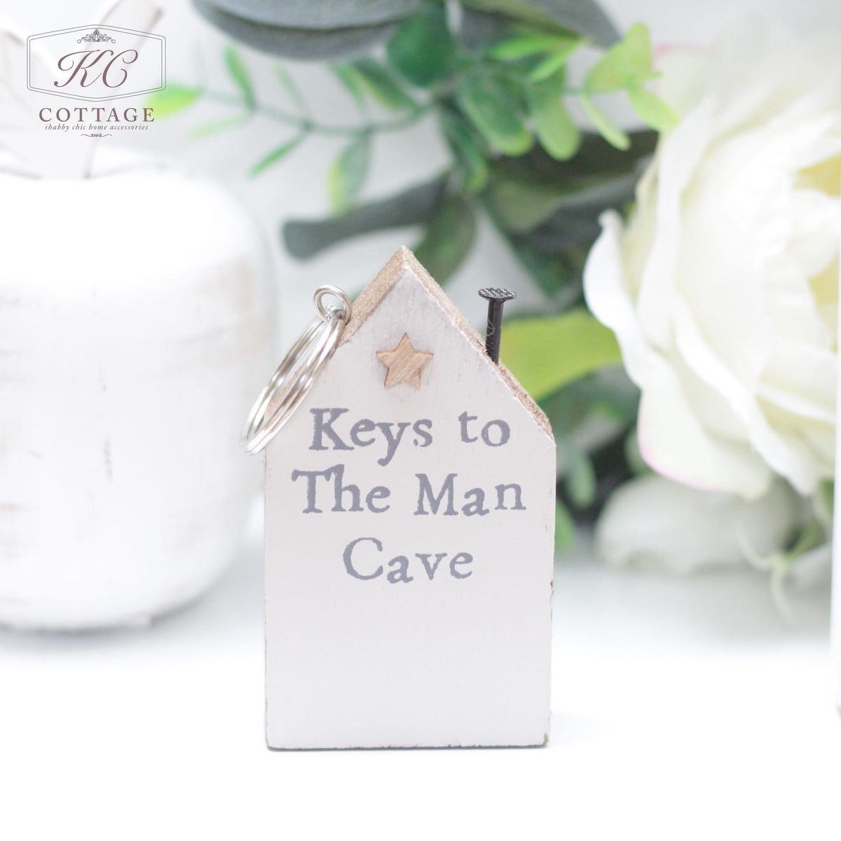 A small Chunky House Wooden Keyring shaped like a house with a star decoration at the top left is perfect for home decor. It has the text "Keys to The Man Cave" written on it. A keyring and nail are attached for hanging keys, set against a background of greenery and a faintly visible white vase.