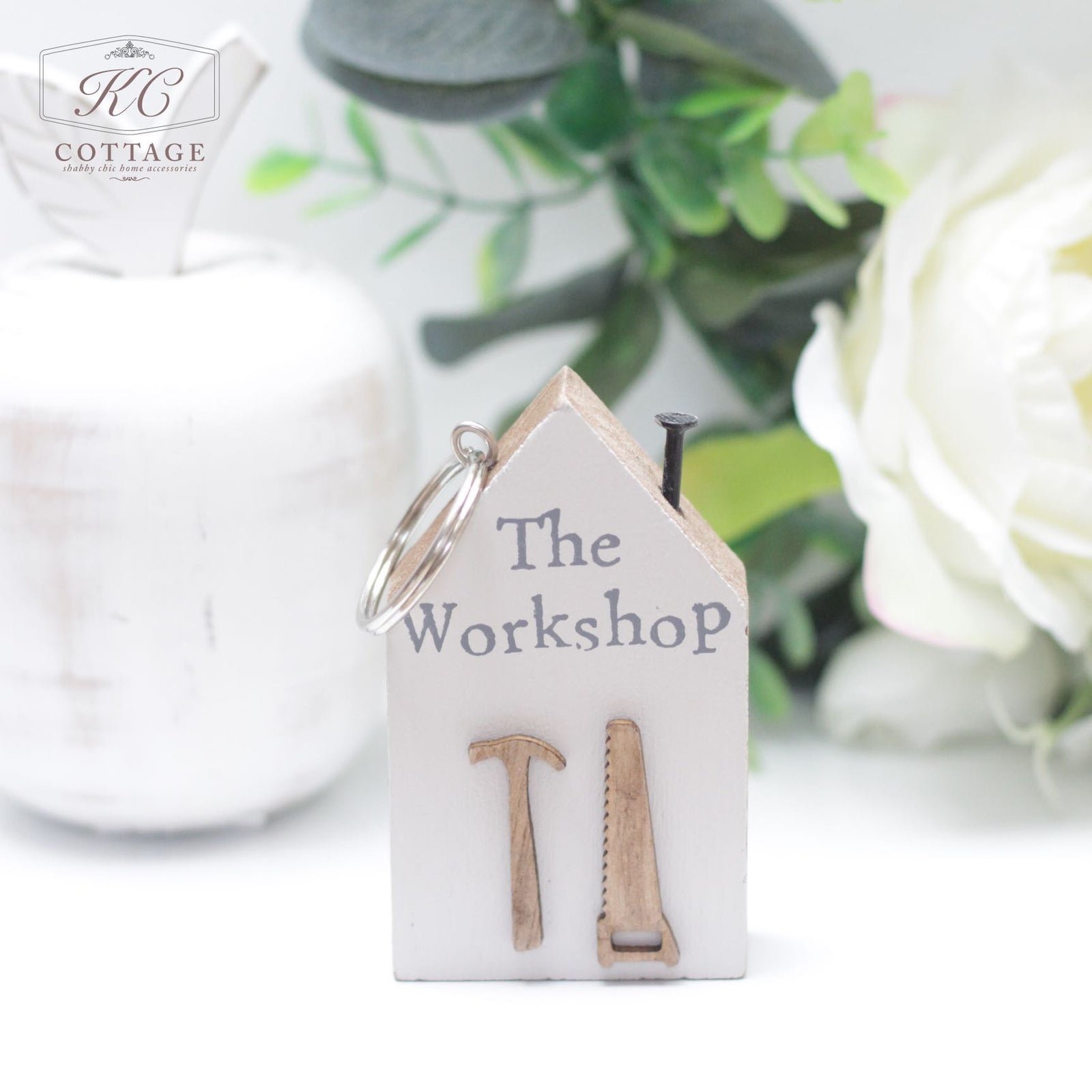 A small, house-shaped key holder with the text "The Workshop" on it, featuring miniature wooden hammer and saw decorations. Perfect for home decor, it's placed on a white surface with a white vase and greenery in the background. The Chunky House Wooden Keyring is attached at the top.