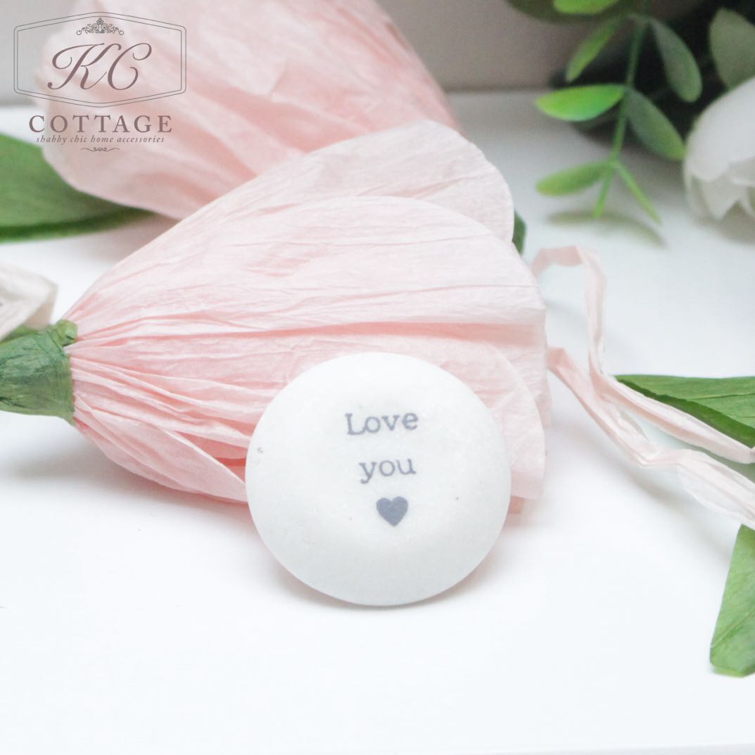 A small, round, white object with the text "Love you" and a heart symbol printed on it is placed in front of pink paper flowers and green leaves. This charming piece adds a lovely touch to any home decor. This Marble Sentiment Tokens in gift bag is sure to warm hearts and bring beauty to your space.