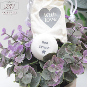 A small decorative bag with "With love" printed on it hangs among green and purple foliage. Nestled in the leaves is a round stone with the words "Best Friend" and a star printed on it. This charming piece, Marble Sentiment Tokens in gift bag, adds a touch of home decor to any space.
