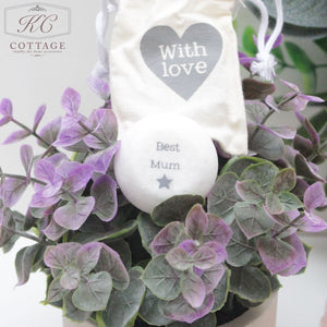 A Marble Sentiment Token in a gift bag with "Best Mum" and a star logo rests on a bed of purple and green leaves in a potted plant, perfect for adding a touch of personal home decor. Hanging above the token is a small white pouch with a heart and the text "With love.