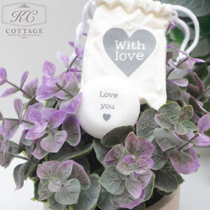 A small white drawstring bag labeled "Marble Sentiment Tokens in gift bag" rests atop a potted plant with purple leaves. In front of the bag is a white round object with the words "Love you" printed on it, accompanied by a small heart. This charming piece adds warmth to any home decor setting.