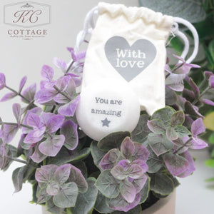 An arrangement of purple flowers with green leaves is pictured. A white drawstring pouch with a heart shape and the words "With love" hangs above the flowers. In the center is a white stone inscribed with "You are amazing" and a small star below the text, perfect for your Marble Sentiment Tokens in gift bag.