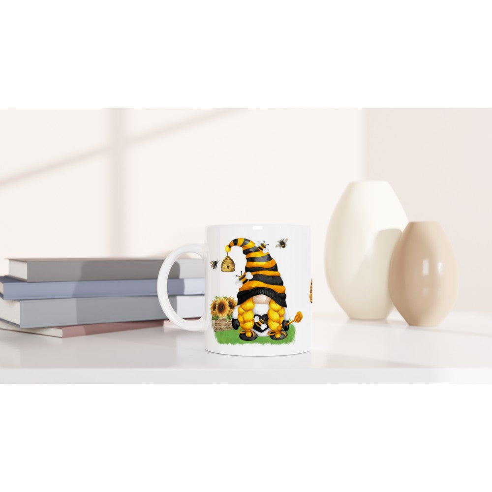 A Bee & Sunflower Gonk Mug featuring an illustration of a gnome dressed as a bee sitting among sunflowers and bees, perfect for adding charm to your home decor. The background includes a stack of books and two vases, one white and one beige, placed on a light-colored surface.