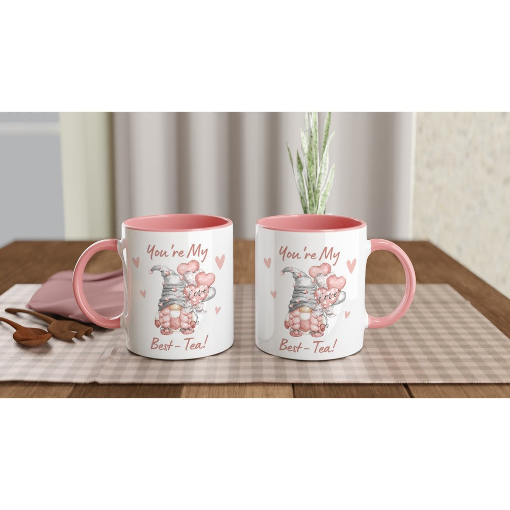 Two white mugs with pink handles and rims sit on a checkered placemat on a wooden table. Each You're My Best-tea Gonk Mug features an image of a cute animal with the text "You're My Best-Tea!" in pink letters, surrounded by small pink hearts, adding a charming touch to your home decor.