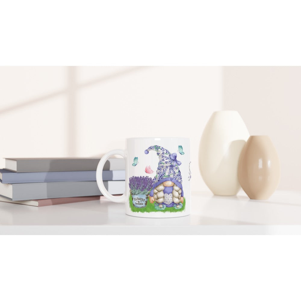 A Gonk Lavender & Butterfly Mug showcases an illustration of a gnome house adorned with flowers and butterflies. It is placed on a light-colored surface next to two stacked books and decorative vases, perfect for any home decor setting, with a soft, shadowed background.